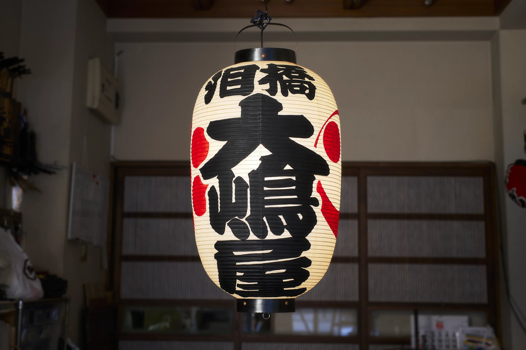 A paper lantern lettered with the Ōshimaya name, lit against a dark wall